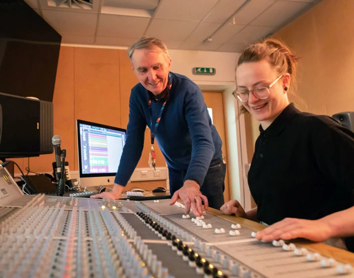 A lecturer and student working together at a large mixing console, discussing adjustments to the sound levels. A lecturer and student working together at a large mixing console, discussing adjustments to the sound levels.